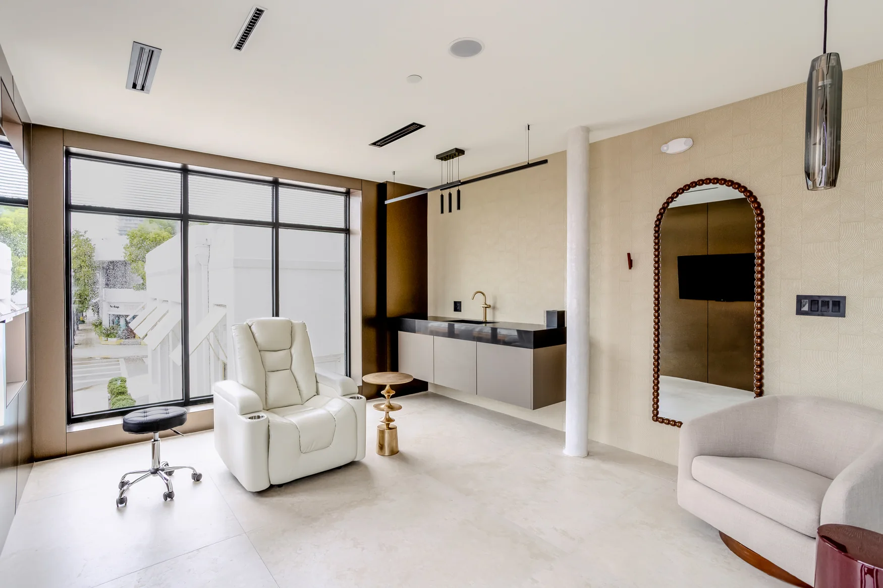 White reclining chair beside a large window, with a sink, mirror and cream sofa.