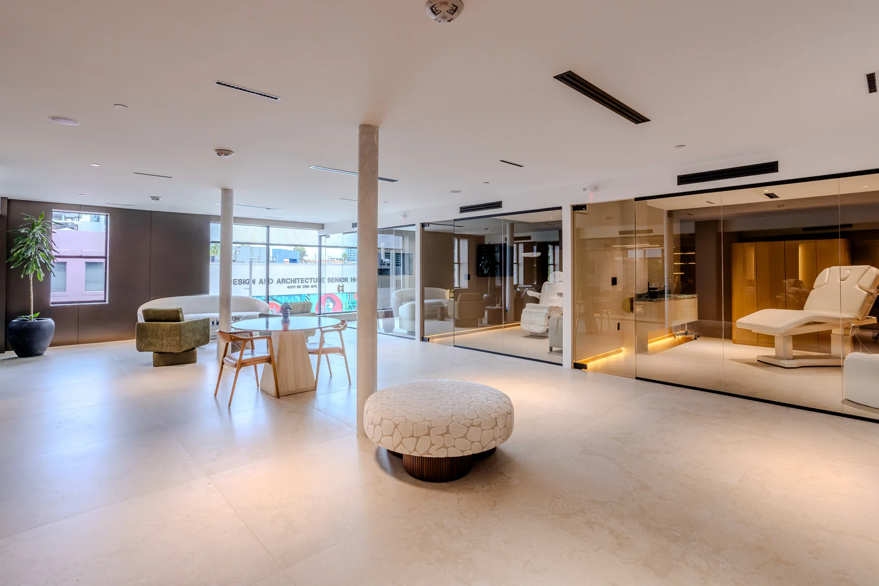 Open lounge with a round table, ottoman, large windows and glass partitions around reclining-chair rooms.