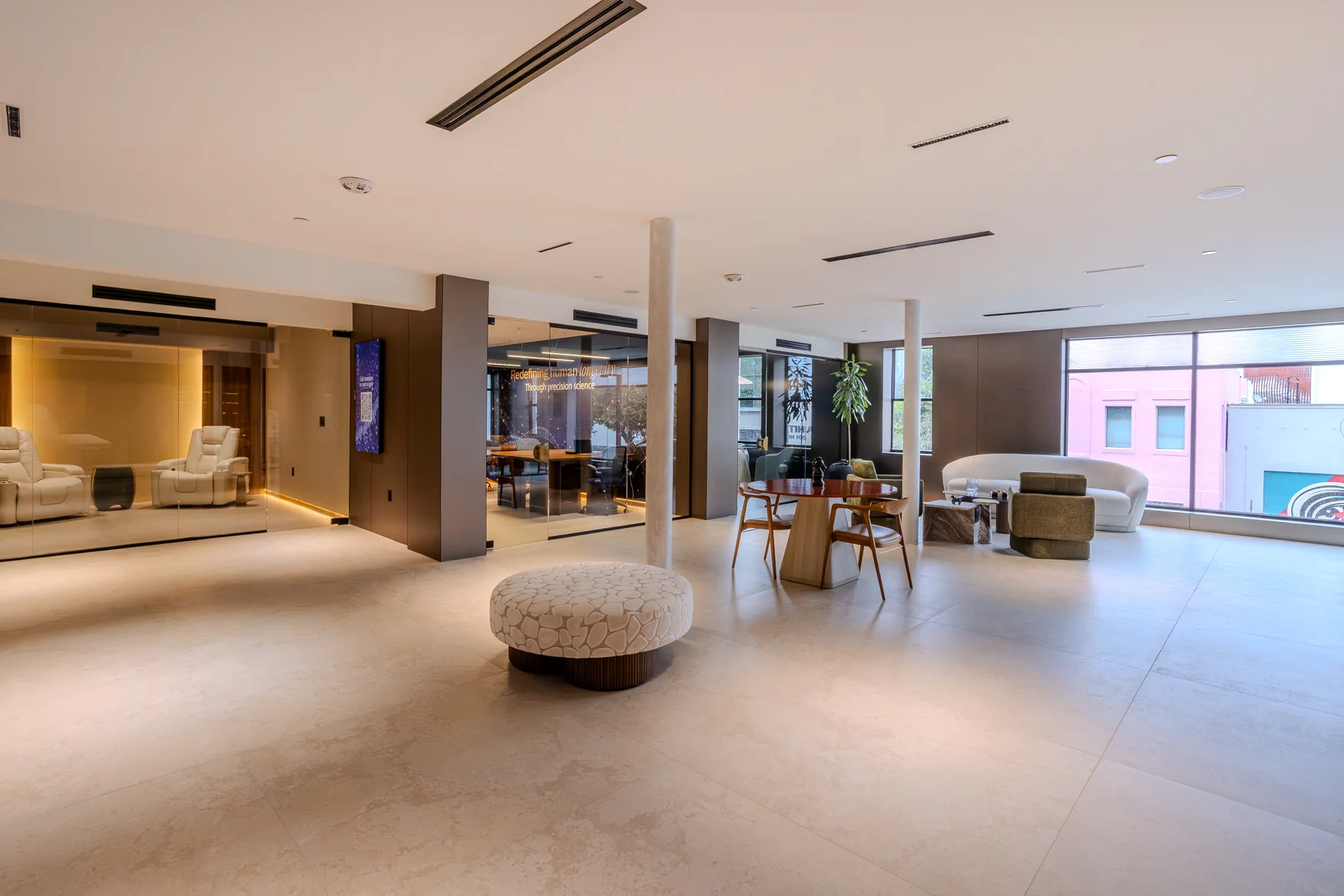 Wide view of the lounge with a pale floor, round seating, a table and glass-fronted rooms.